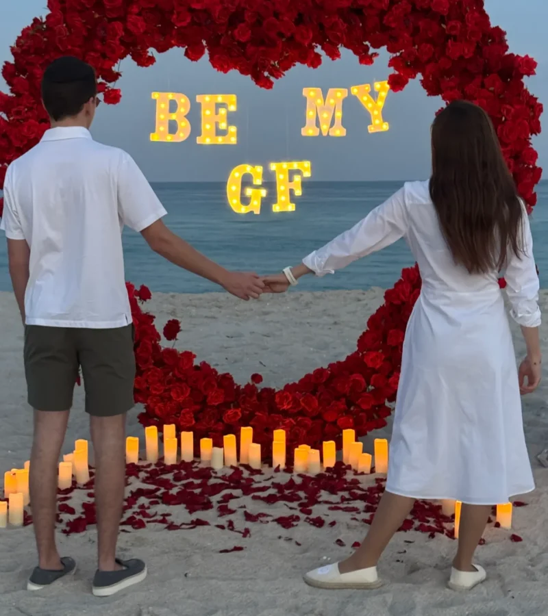 Couple holding hands in front of "Be My GF" sign in red rose arch